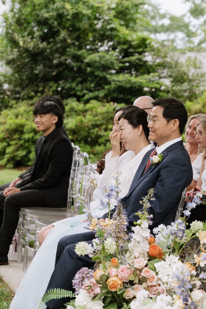 Family members seated during an outdoor wedding ceremony surrounded by colorful floral arrangements.