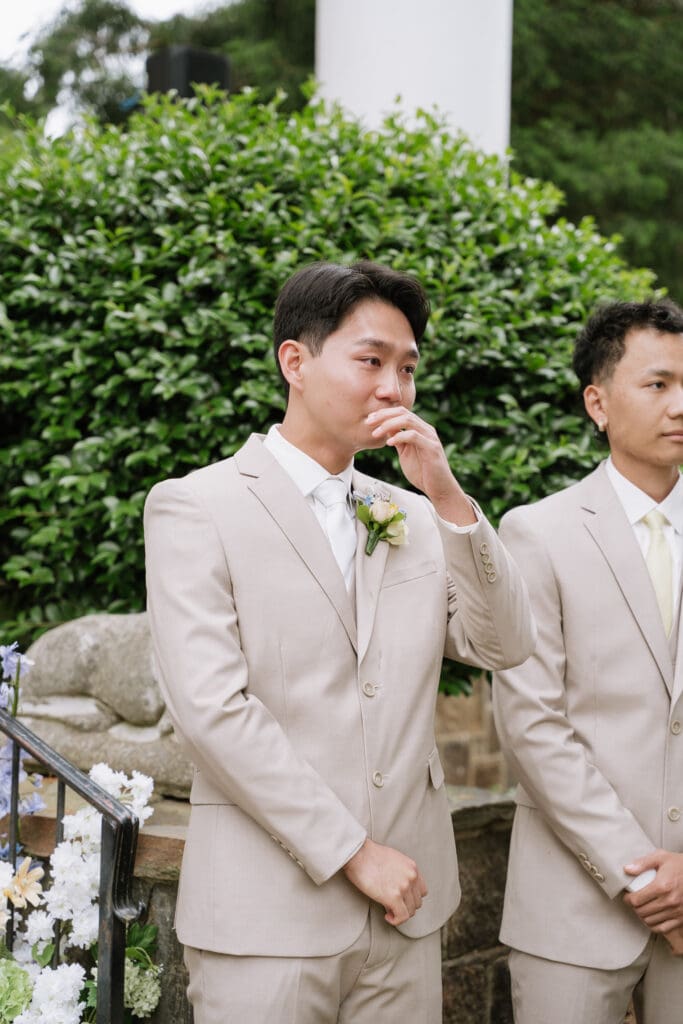 Groom becoming emotional while standing beside his groomsmen during an outdoor wedding ceremony.