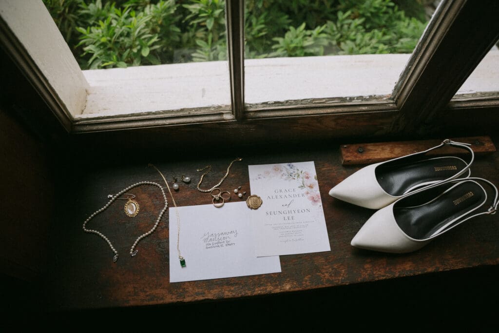 Wedding details including invitations, jewelry, and bridal shoes arranged beside a window.