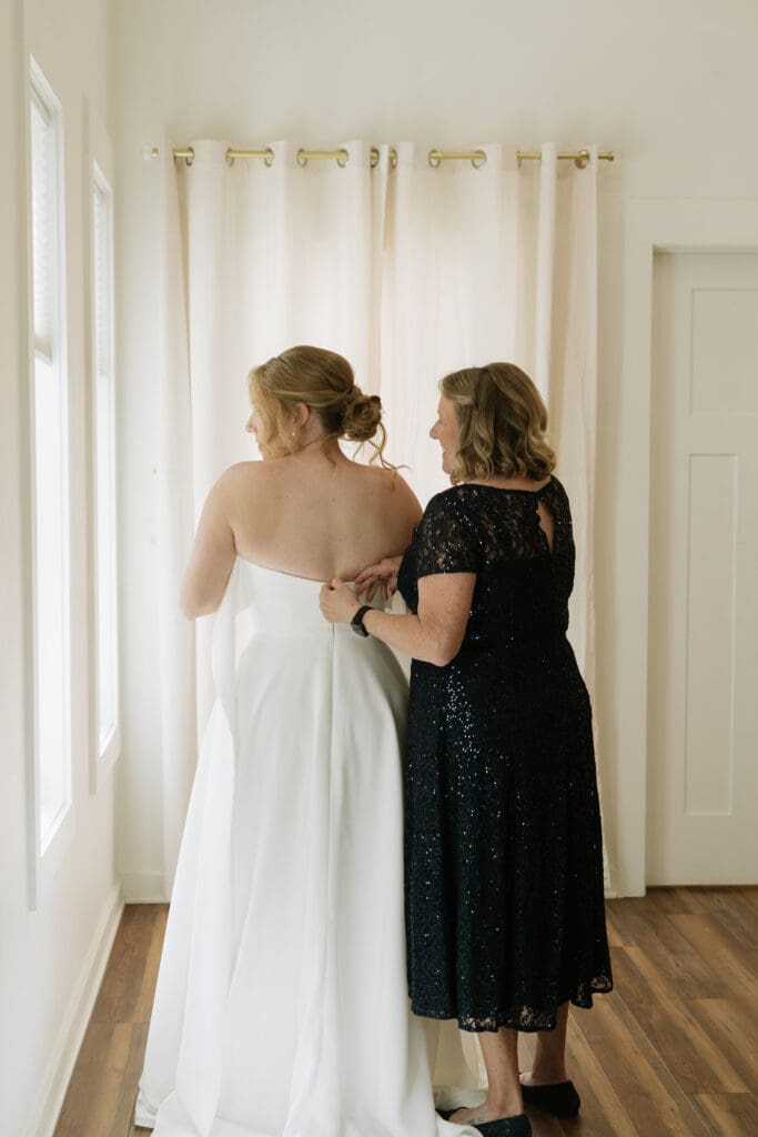 Mother helping her daughter get ready by fastening her wedding dress before the ceremony.