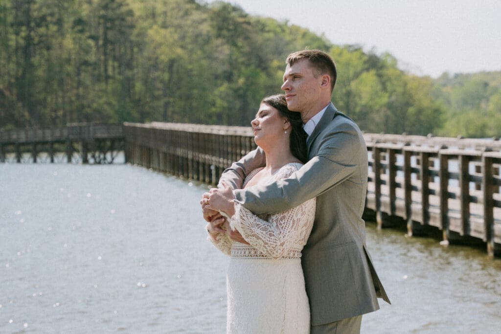 Bride and groom embracing beside the water at an outdoor wedding venue with natural scenery and colorful surroundings.